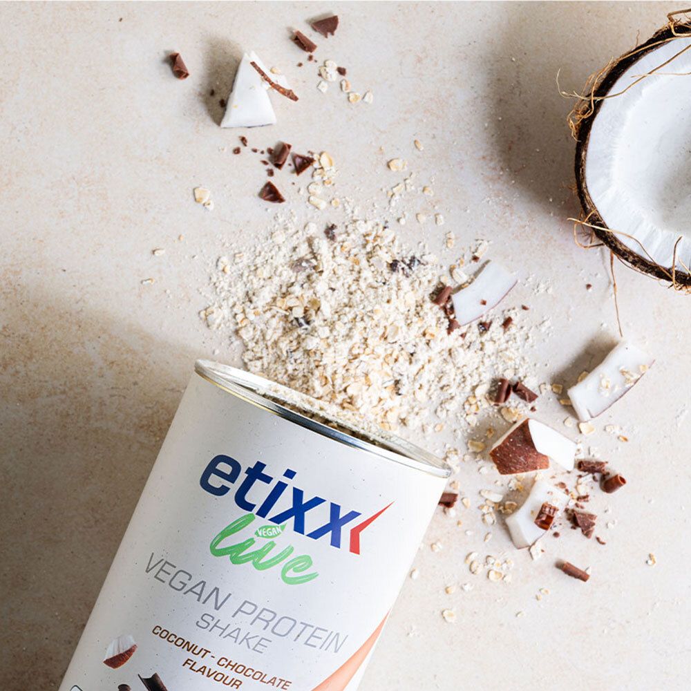 Boîte ouverte de etixx Live Vegan Protein Shake. Morceaux de noix de coco et de chocolat dispersés. Moitié de noix de coco en arrière-plan.