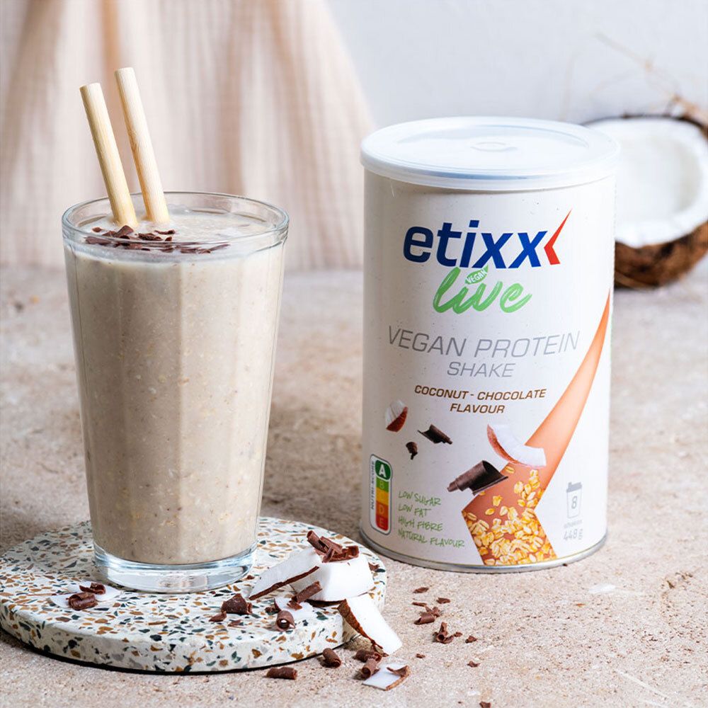 Verre de etixx Live Vegan Protein Shake. Garniture noix de coco et chocolat. Boîte à côté. Pailles. Moitié de noix de coco.