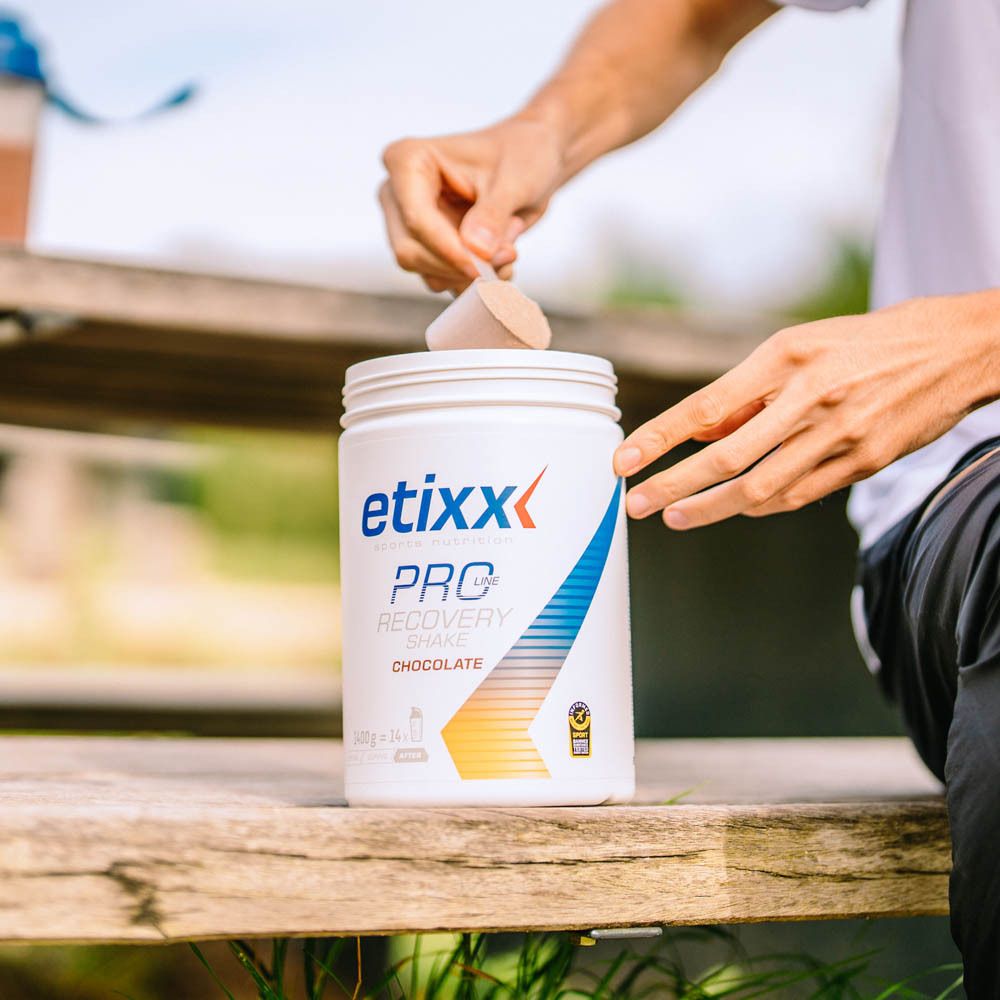 Persoon giet poeder in een pot. Opschrift: etixx, PRO, Recovery Shake Chocolate. Op houten bank.