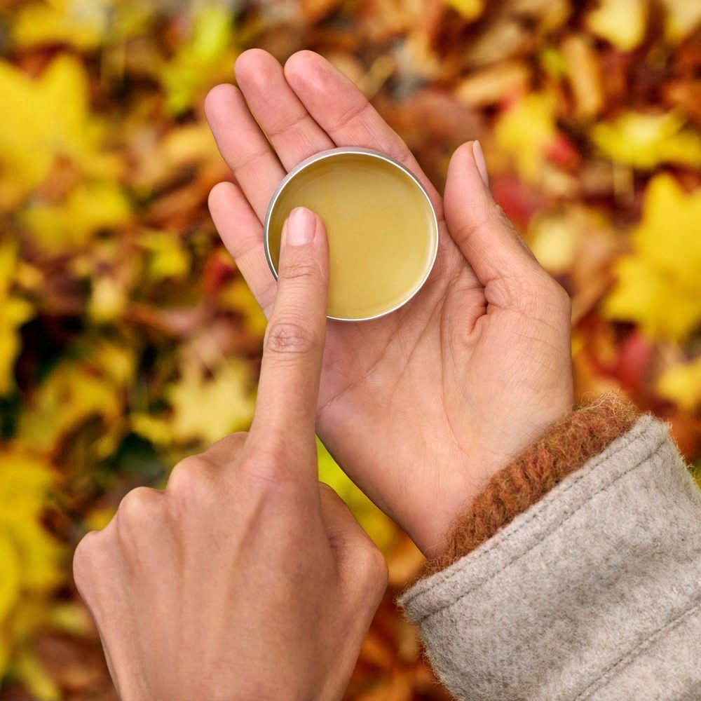 Mains tenant un pot ouvert de baume jaune. Un doigt touche le baume. Fond: feuilles d'automne.