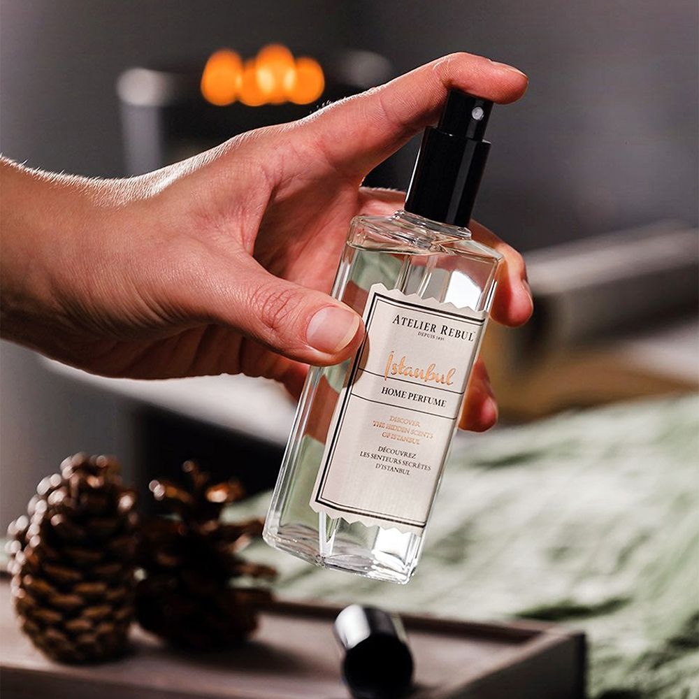 Hand hält ATELIER REBUL Istanbul Home Parfüm-Flasche. Rechteckige Flasche mit schwarzem Sprühkopf. Hintergrund unscharf.