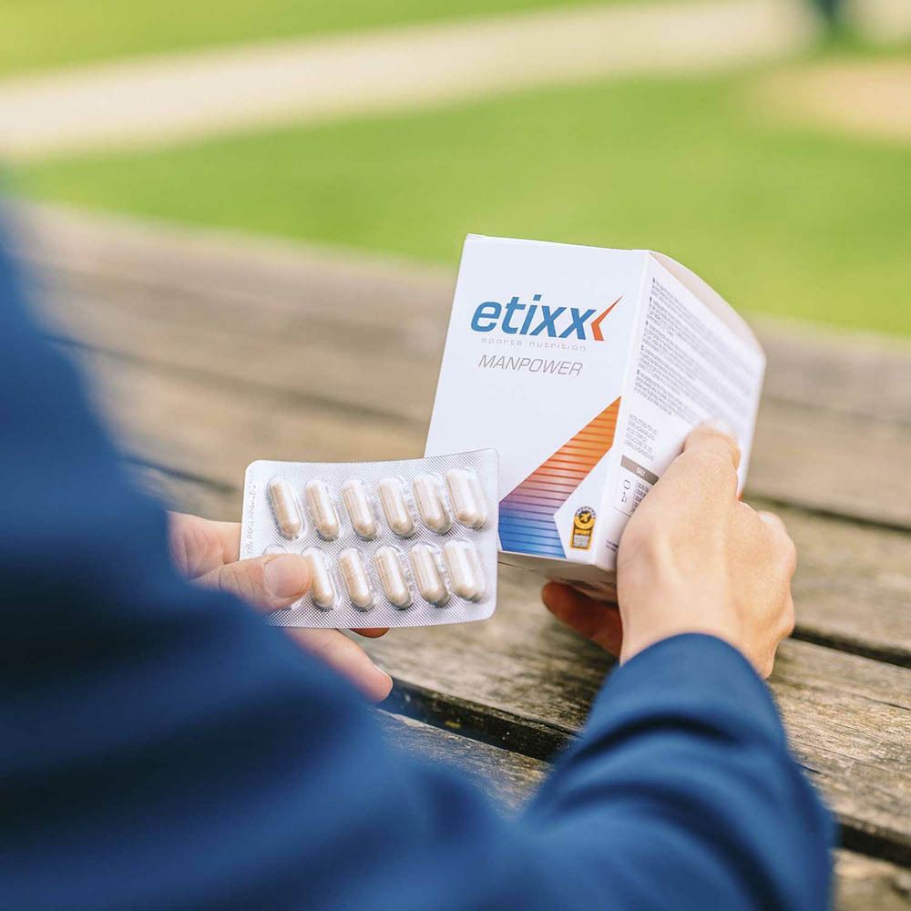 Persoon houdt een doos en blisterverpakking met capsules vast. "etixx MANPOWER" doos. Buiten, vage achtergrond.