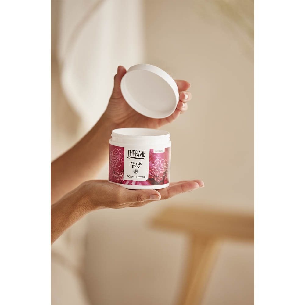Persoon houdt een witte pot met rode banderol vast. Opschrift THERME, Mystic Rose, Body Butter. Deksel open.