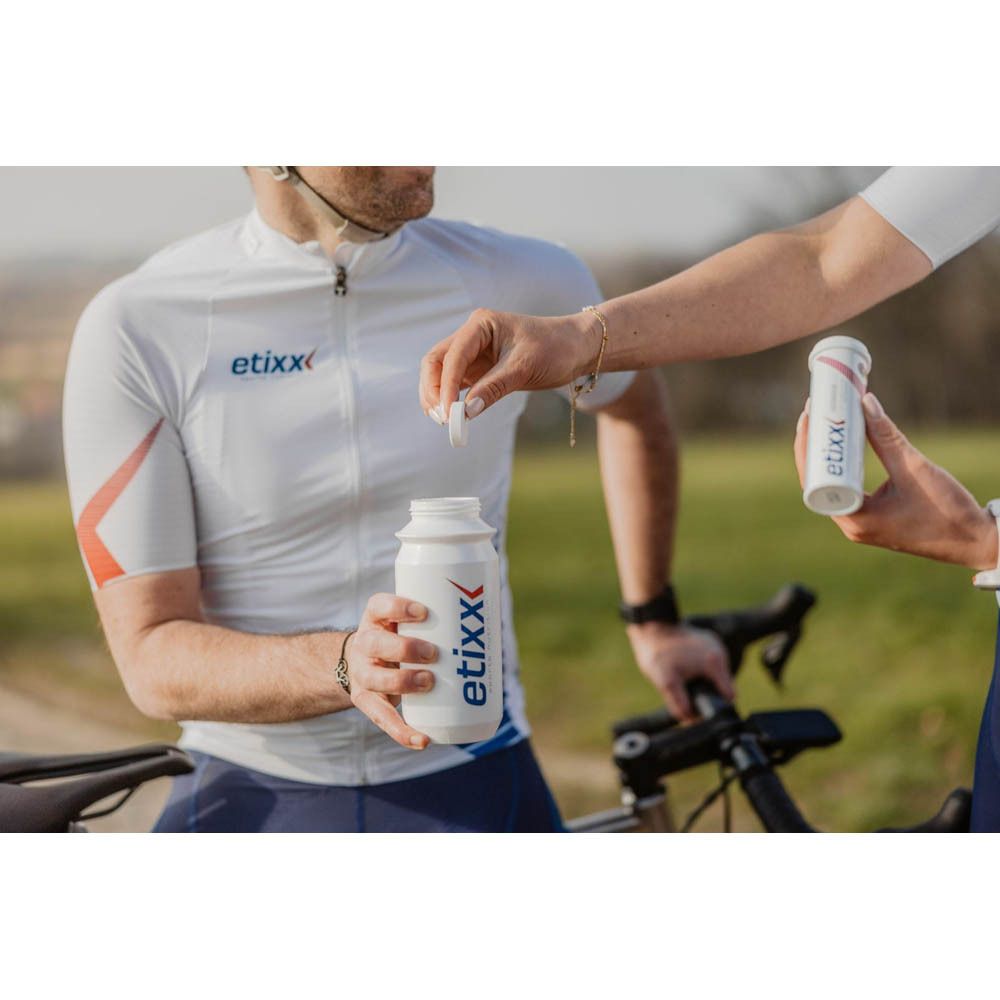Twee personen buiten. Eén persoon houdt een witte fles met Etixx-logo vast, de ander een tablet. Op de achtergrond een fietser.