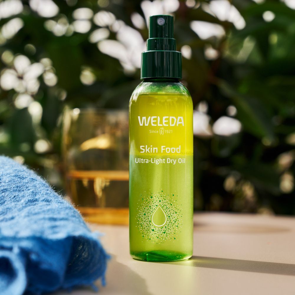 Grüne Sprühflasche mit WELEDA-Logo und Skin Food-Aufschrift. Steht neben einem Glas und einem blauen Tuch.