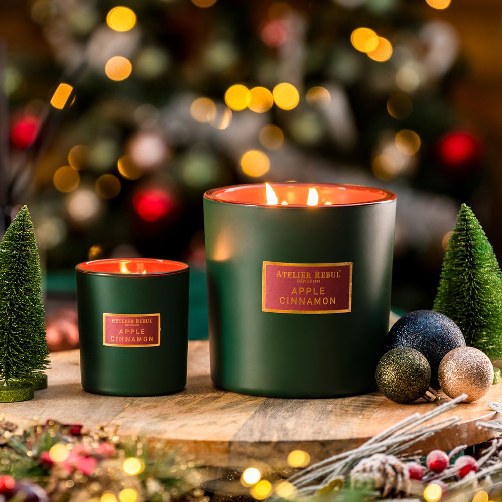 Deux bougies, une grande, une petite, avec étiquettes "ATELIER REBUL Apple Cinnamon". Décorations de Noël en arrière-plan.