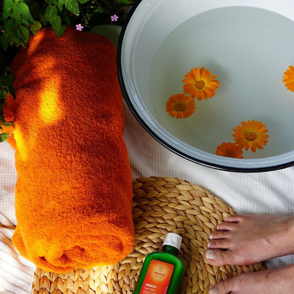 Füße in einer Schüssel mit Wasser und Blüten. Orangefarbenes Handtuch, Flasche mit Produktetikett und Korbuntersetzer.