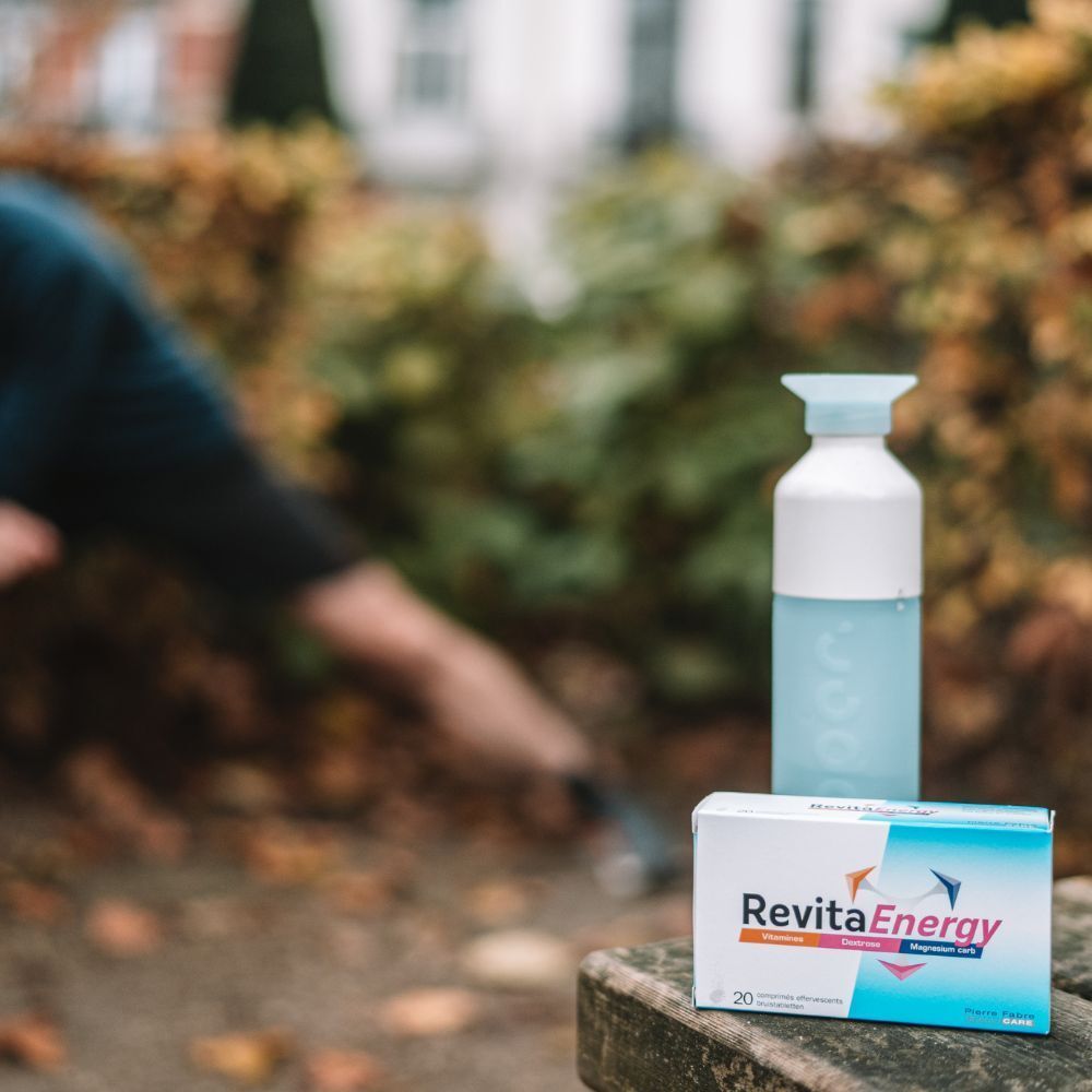 REVITA ENERGIE-Schachtel und Wasserflasche auf einer Holzbank. Im Hintergrund eine Person beim Training.