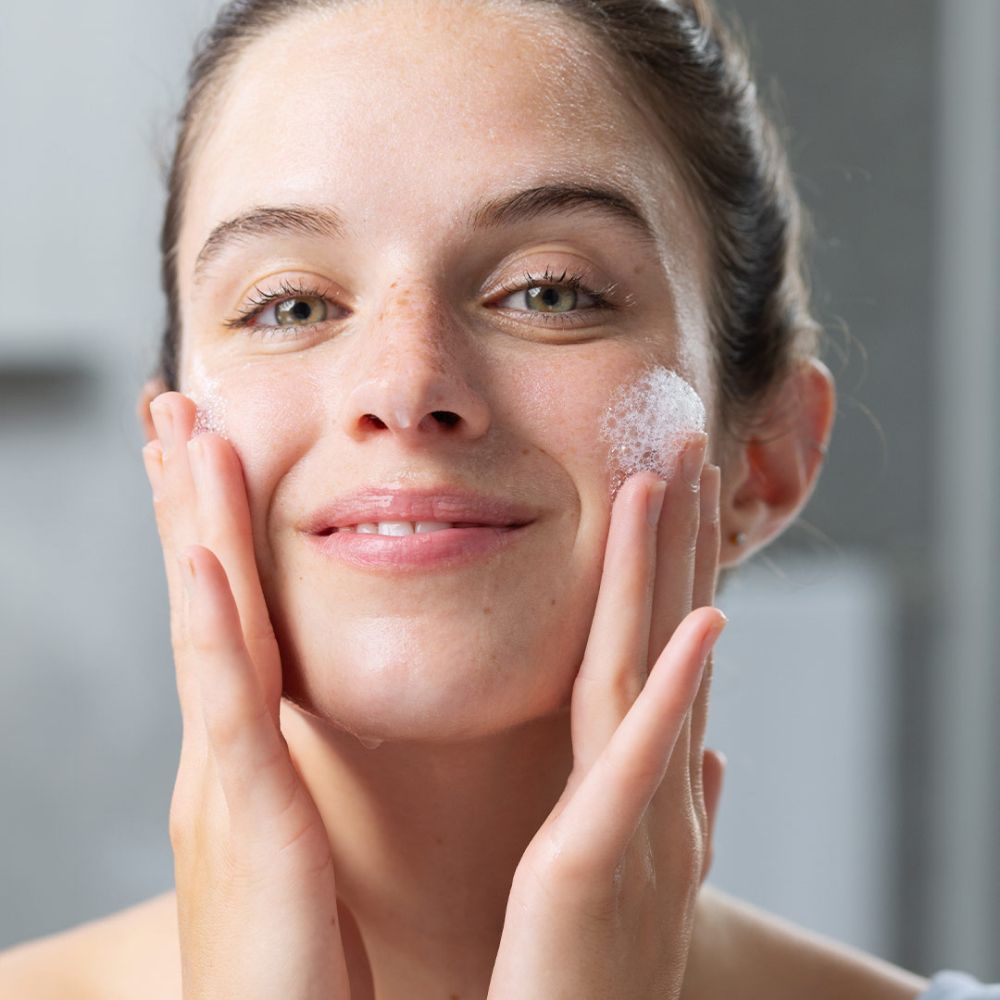Femme avec de la mousse sur le visage. Elle touche son visage avec ses mains. Sourit.