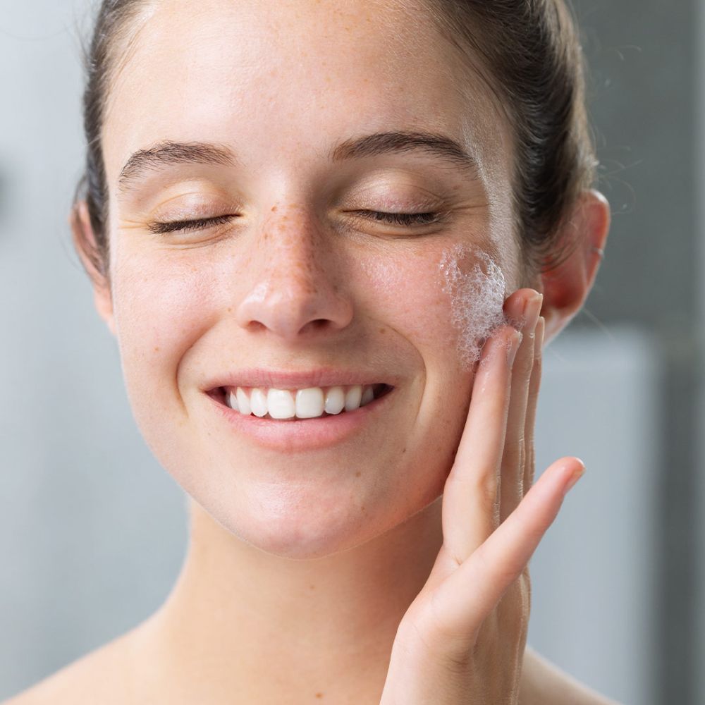 Femme aux yeux fermés avec de la mousse sur le visage. Elle touche son visage avec ses mains.