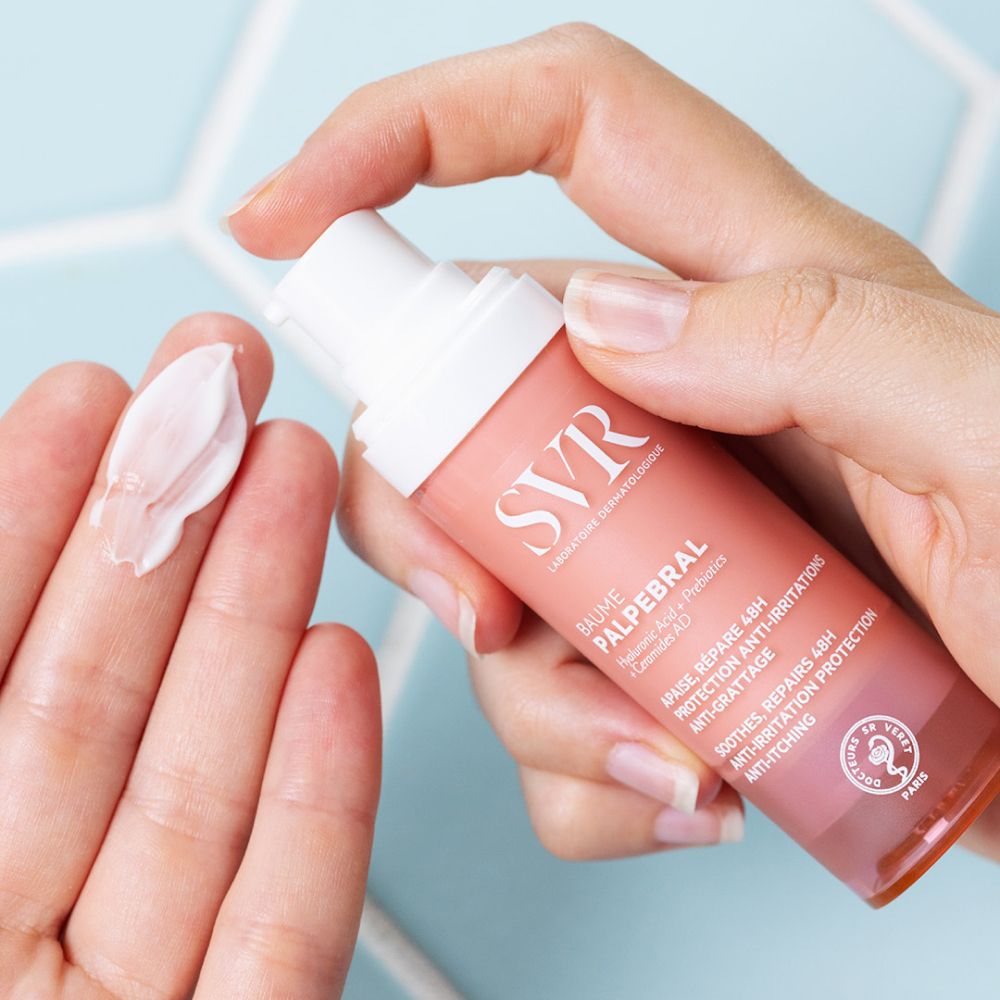 Hand met SVR Baume Palpébral. Crème op vinger. Roze en witte fles. Tekst: Baume Palpébral.