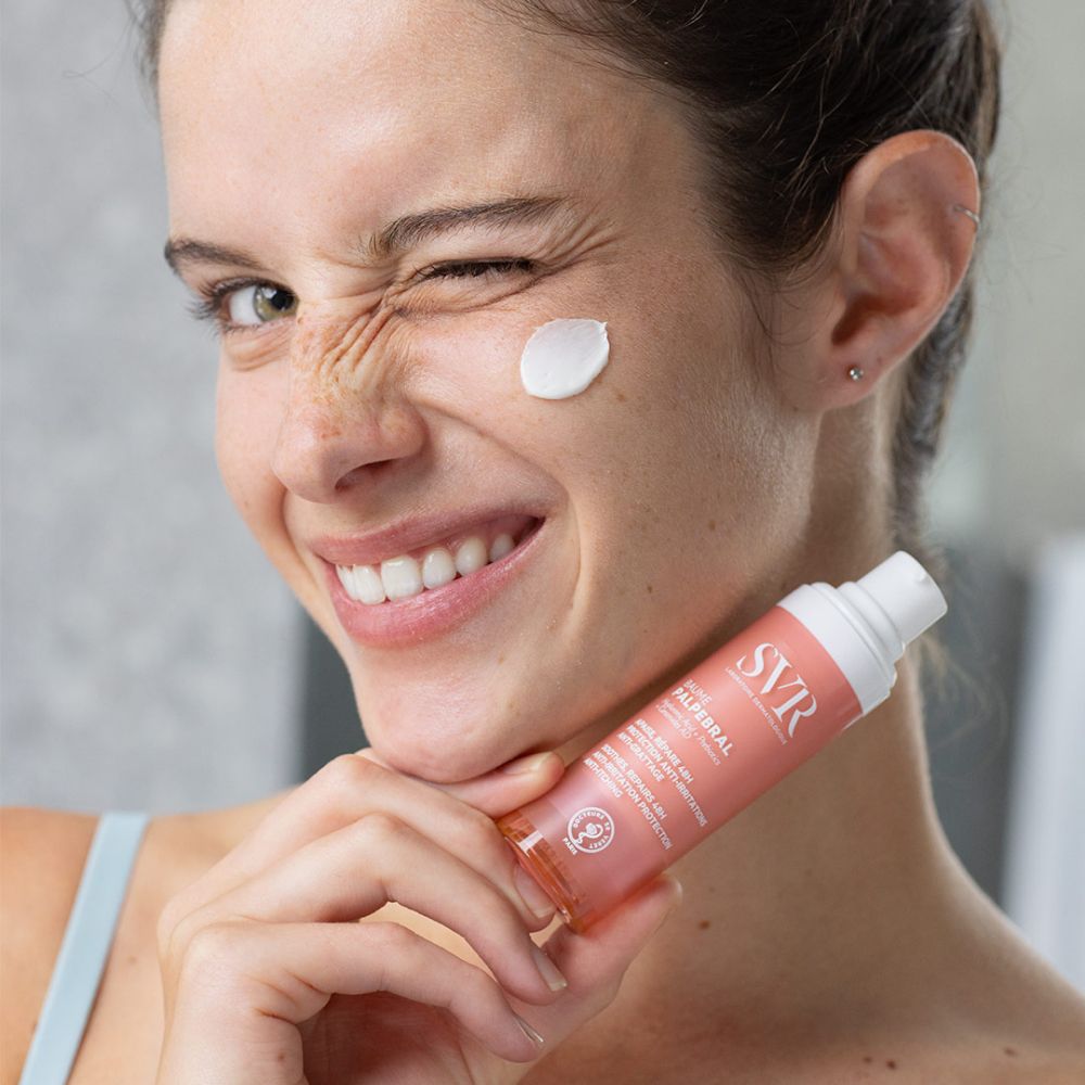Vrouw brengt SVR Baume Palpébral aan. Roze en witte fles. Tekst: Baume Palpébral. Crème op huid.