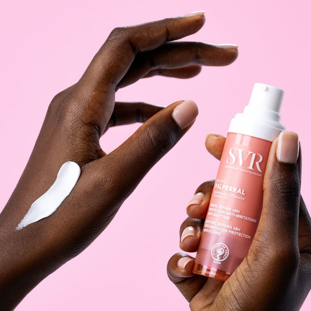 Hand met SVR Baume Palpébral. Crème op huid. Roze en witte fles. Tekst: Baume Palpébral.