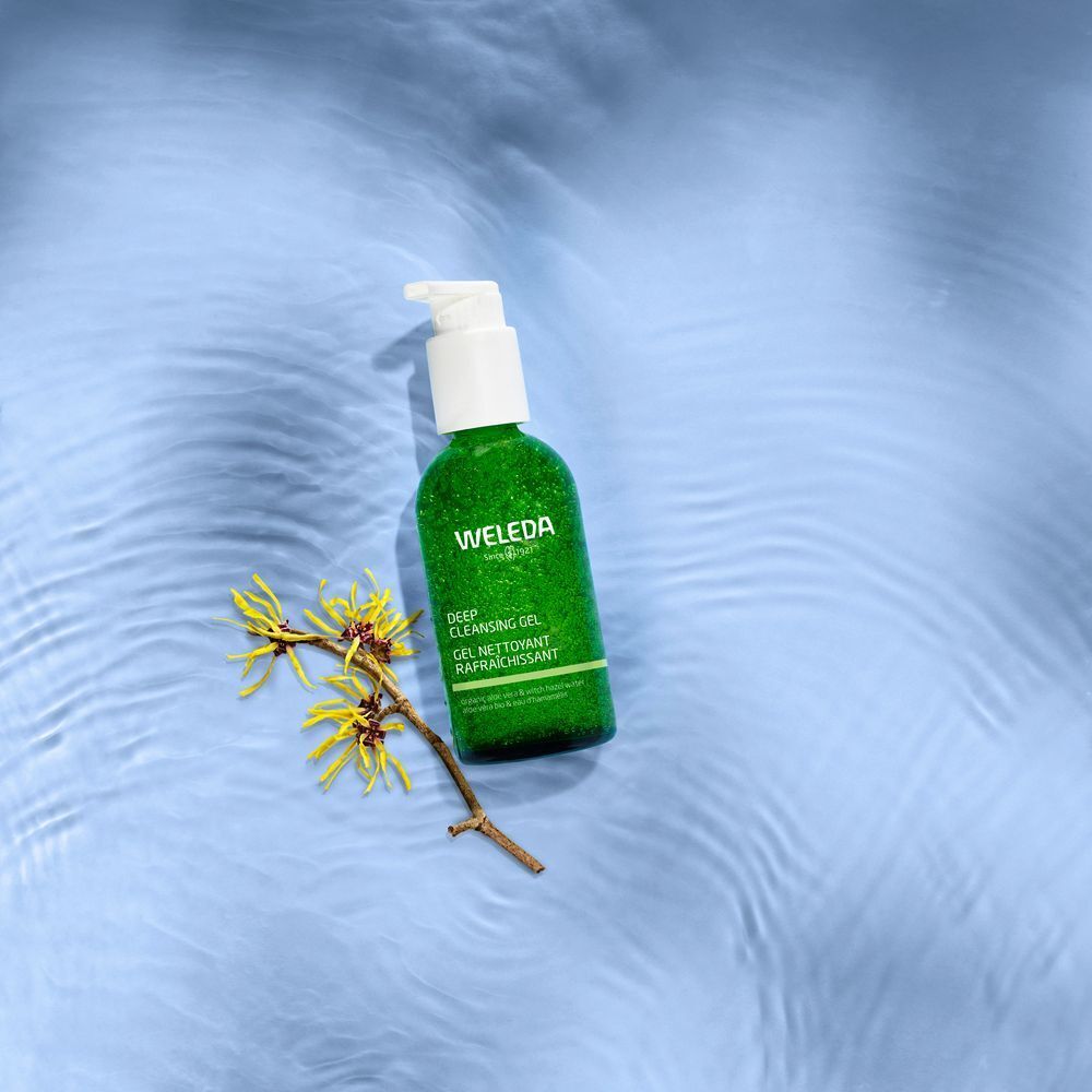 Flacon vert avec pompe blanche sur eau bleue. Inscription: WELEDA, GEL NETTOYANT RAFRAÎCHISSANT. Contient de l'hamamélis. Décor floral.