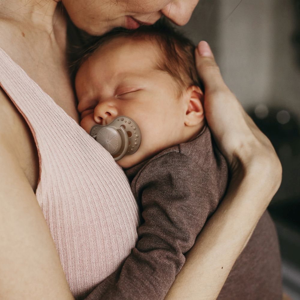 Bébé avec sucette en bouche, tenu par une personne. Sucette aux tons bruns.