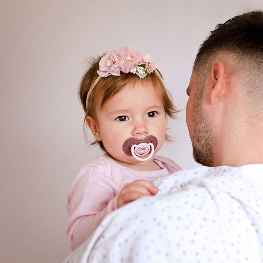 Bébé avec une sucette dans la bouche, tenu par une personne. La sucette est rose.