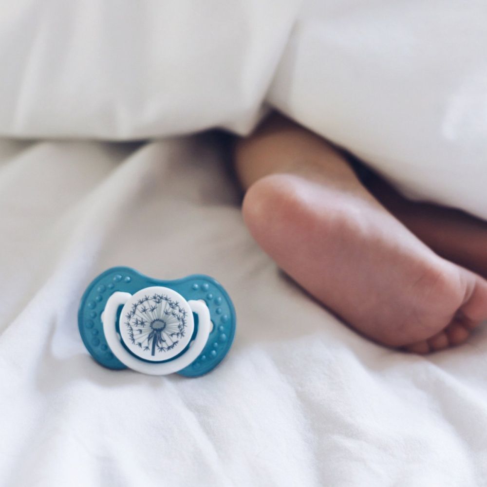 Une sucette bleue avec motif pissenlit à côté d'un pied de bébé sous une couverture blanche.