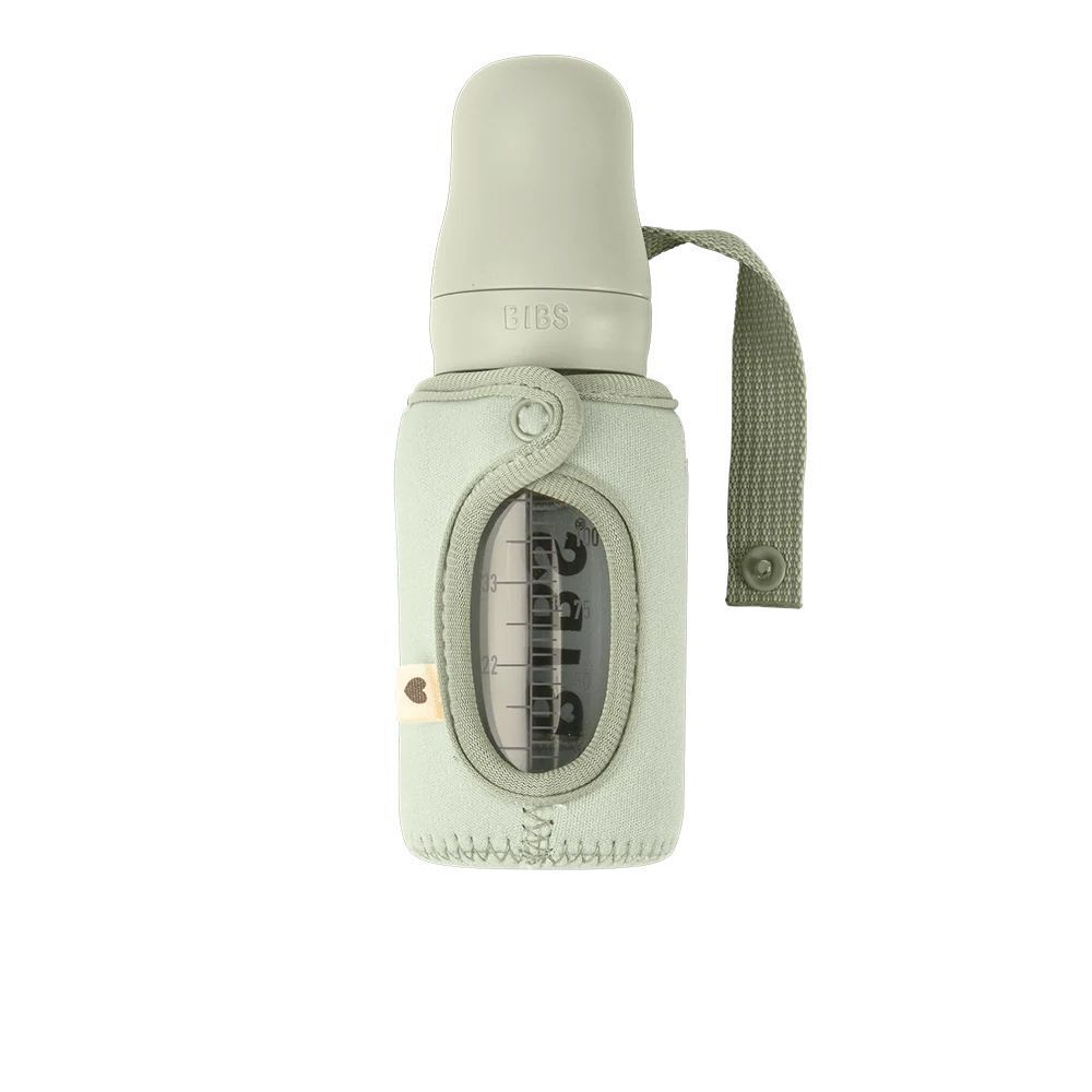 Biberon vert sauge dans un porte-biberon. Échelle visible. Le support a une boucle de transport et une fermeture à bouton-pression.