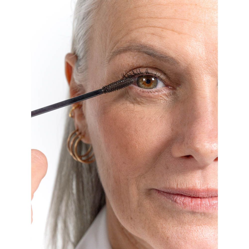 Femme avec applicateur de mascara sur l'œil. Cheveux gris, boucles d'oreilles dorées.