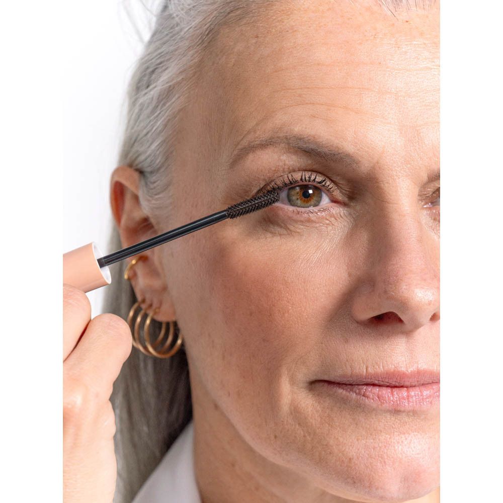 Femme avec applicateur de mascara sur l'œil. Cheveux gris, boucles d'oreilles dorées.