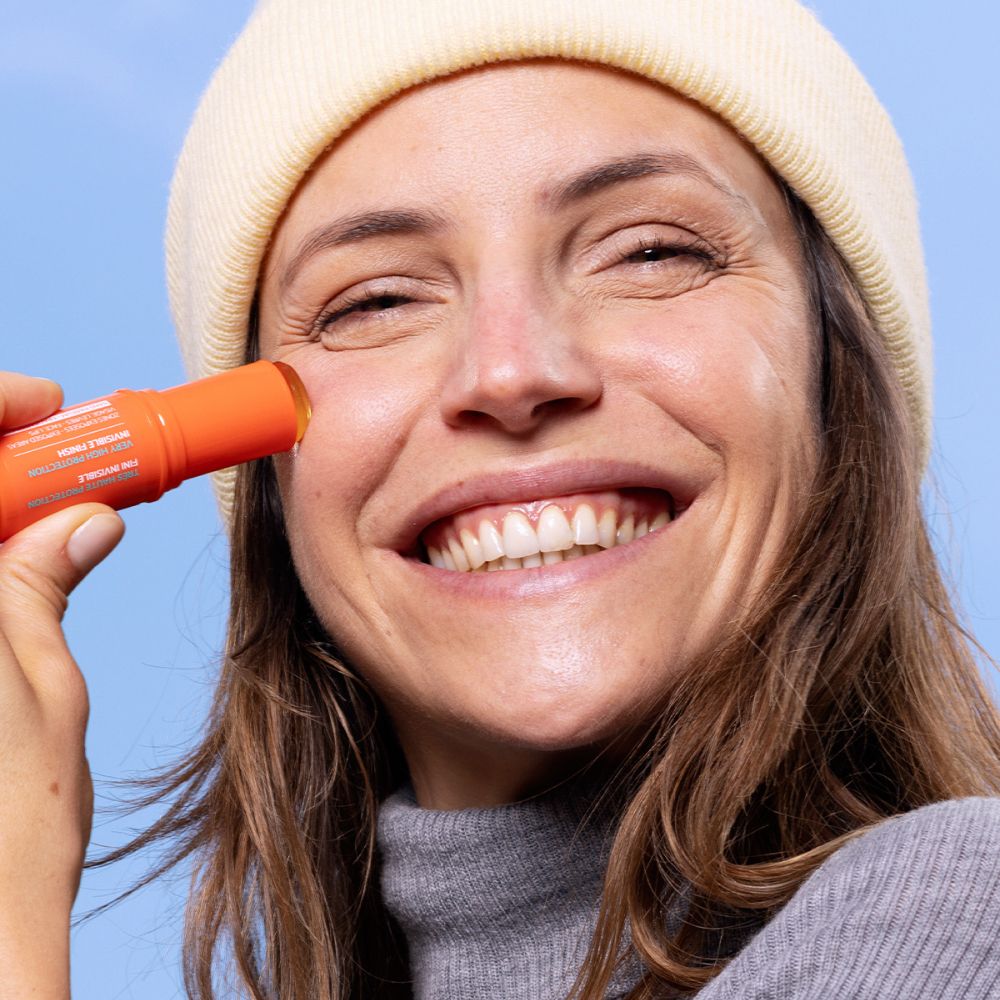 Femme avec bonnet jaune appliquant SVR Easy Stick SPF50+ sur la joue.