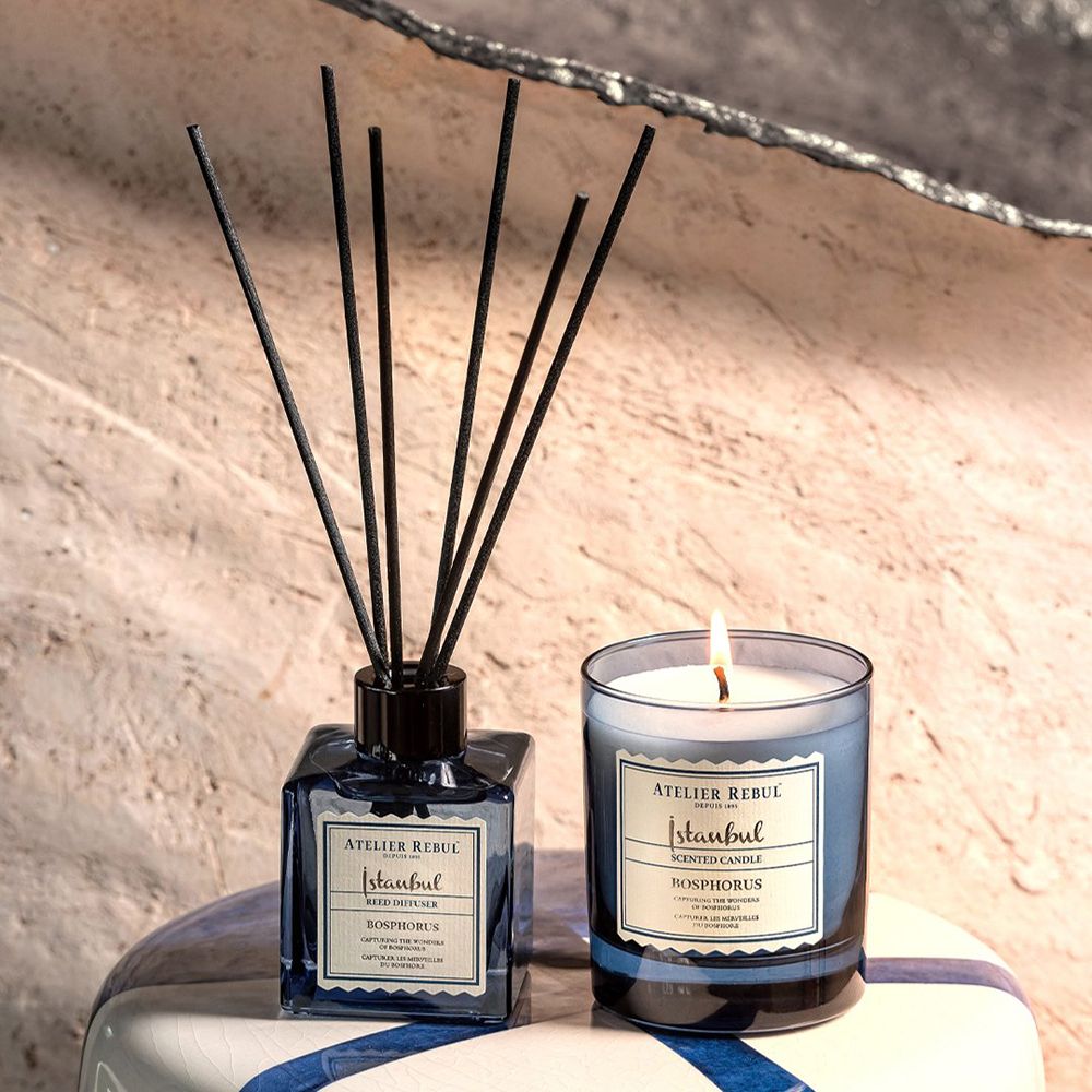 Geurkaars en diffuser. Kaars met vlam, blauw glas. Diffuser met zwarte stokjes, blauw glas. Op tafel.