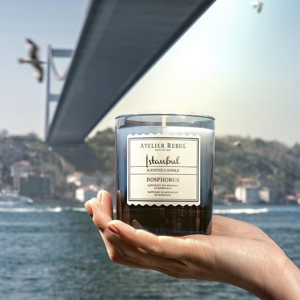 Hand houdt kaars vast. Kaars in blauw glas, wit etiket. Achtergrond: brug, zee, lucht.