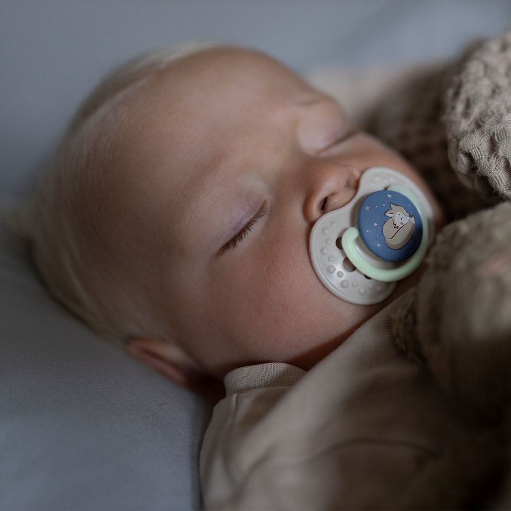 Baby schläft mit Schnuller im Mund. Schnuller mit Fuchs-Motiv. Helles Zimmer, Baby liegt auf Decke.