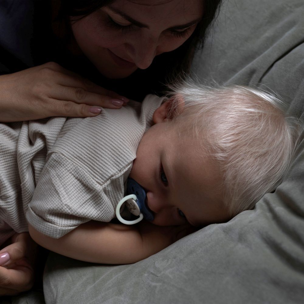 Baby liegt auf einer Couch, Schnuller im Mund. Mutter berührt das Baby. Schnuller blau.