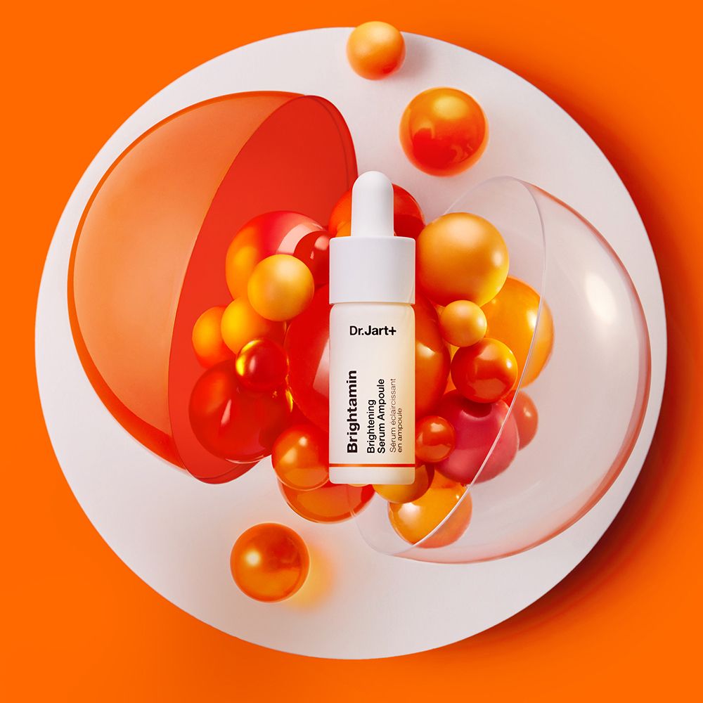 Kleine Flasche mit Pipette auf einer weißen Platte mit orangefarbenen Kugeln. Aufschrift: Dr.Jart+ Brightamin Brightening Serum Ampoule.