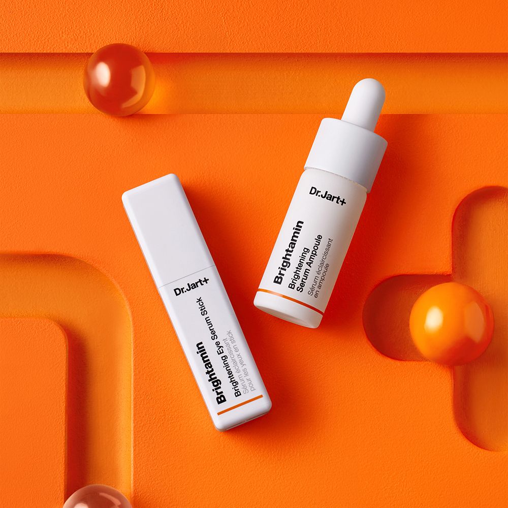 Zwei weiße Tuben auf orangefarbenem Hintergrund. Aufschrift: Dr.Jart+ Brightamin Brightening Eye Serum Stick und Ampoule.