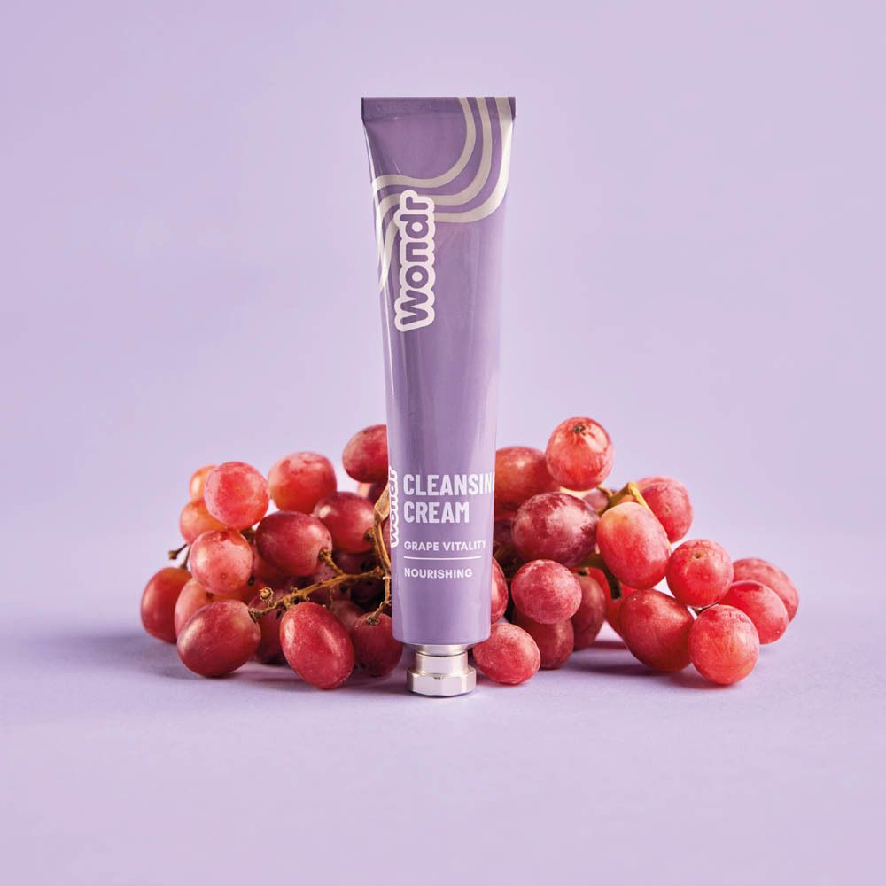 Paarse tube met "Wondr" logo en "Cleansing Cream Grape Vitality" tekst. Ligt op druiven. Op paarse achtergrond.