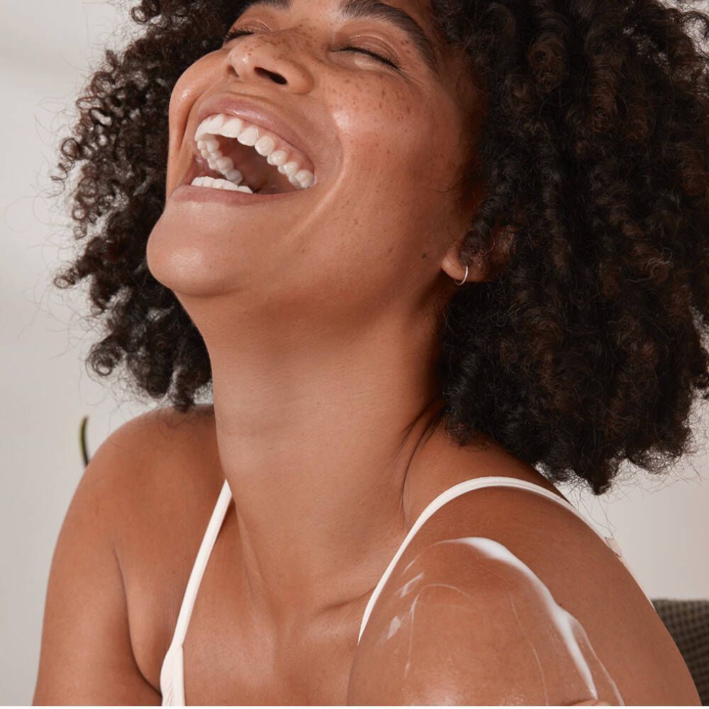 Une femme rit, appliquant une crème sur son bras. Crème blanche visible.