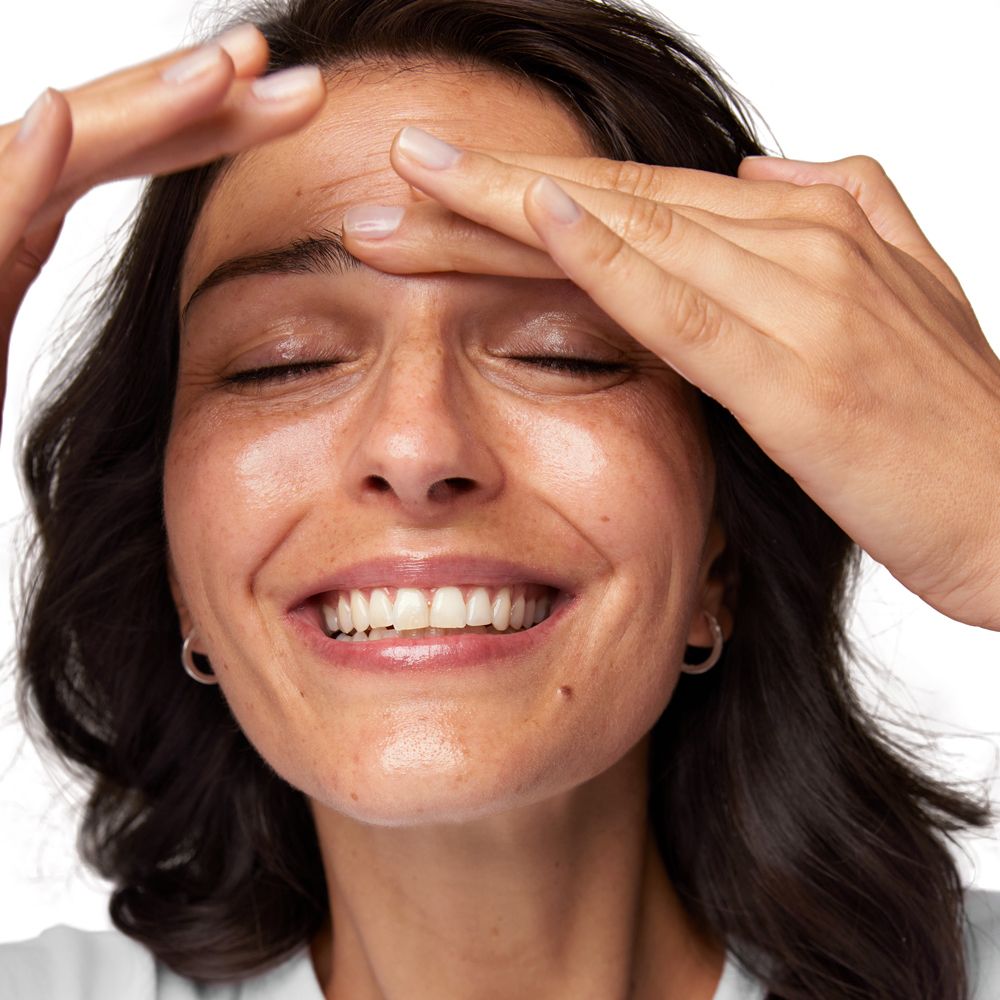 Femme aux yeux fermés, mains sur le front. Peau avec des taches de rousseur. Sourire.