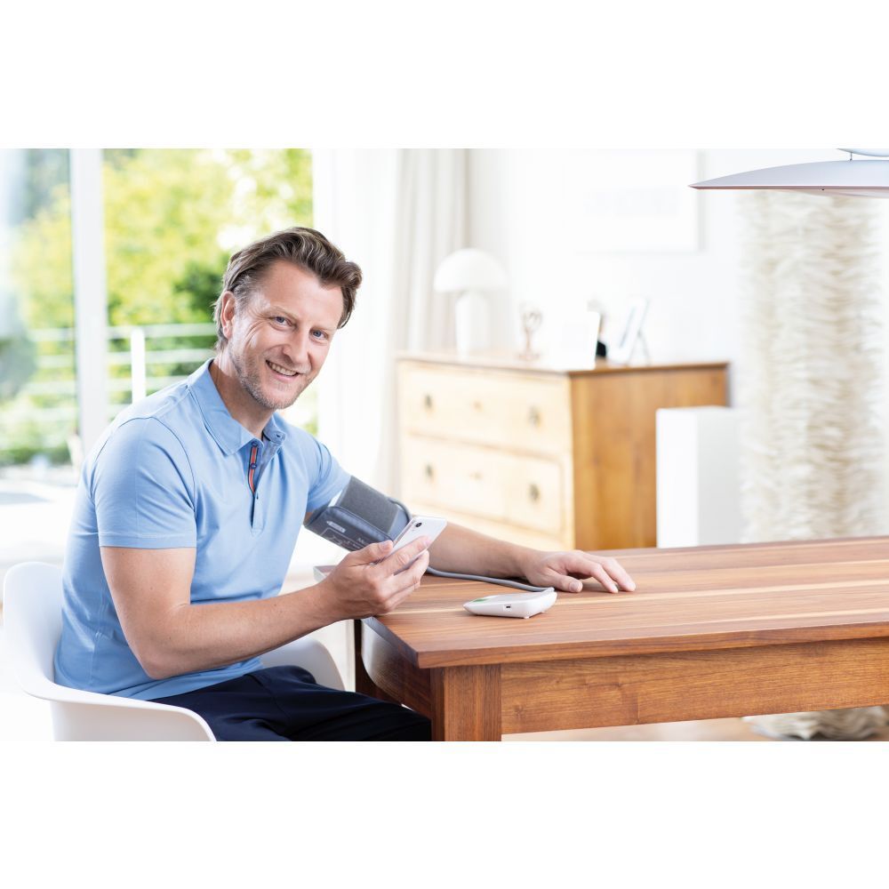 Man zit aan tafel en meet bloeddruk. Bloeddrukmeter en manchet zichtbaar. Merk: medisana.