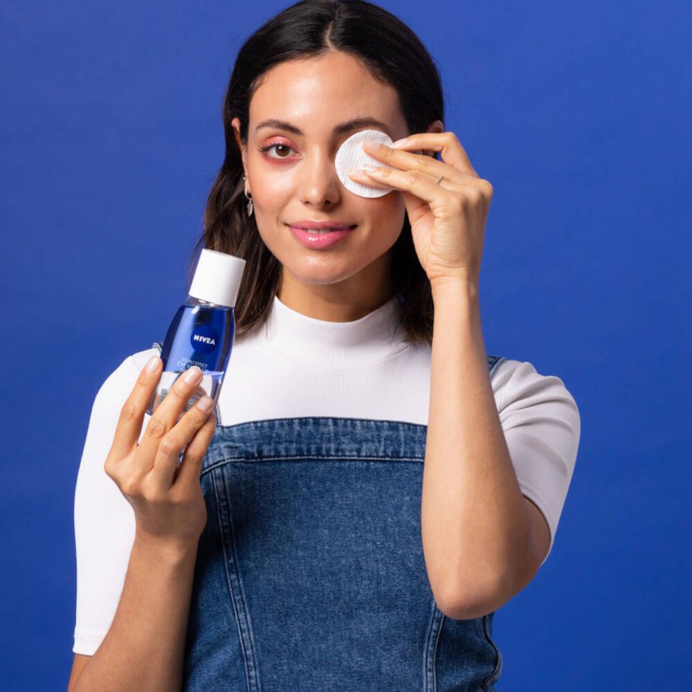 Frau hält NIVEA Augen-Make-up-Entferner und Wattepad. Blaue Wand im Hintergrund. Frau trägt Jeans-Latzhose.
