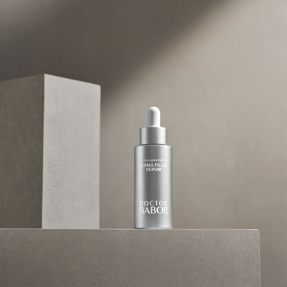 Flacon argenté avec compte-gouttes blanc. Inscription : Derma Filler Serum, Doctor BABOR. Sur surface grise.