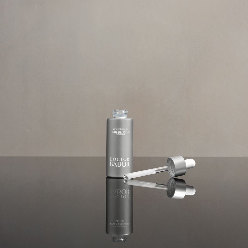 Silberne Flasche mit Tropfer auf glänzender Oberfläche. Aufschrift: Pore Refining Serum. Marke: Doctor Babor.