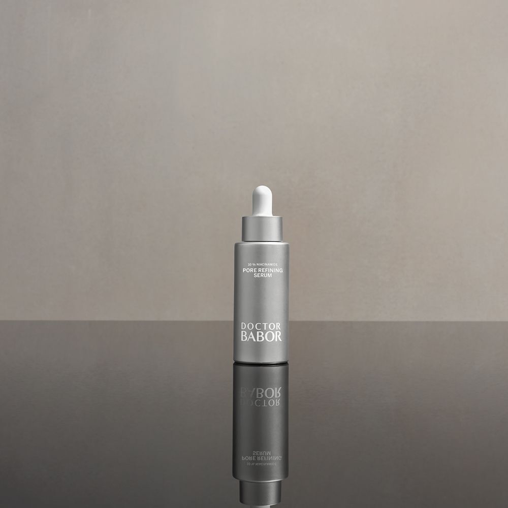 Silberne Flasche mit Tropfer auf glänzender Oberfläche. Aufschrift: Pore Refining Serum. Marke: Doctor Babor.