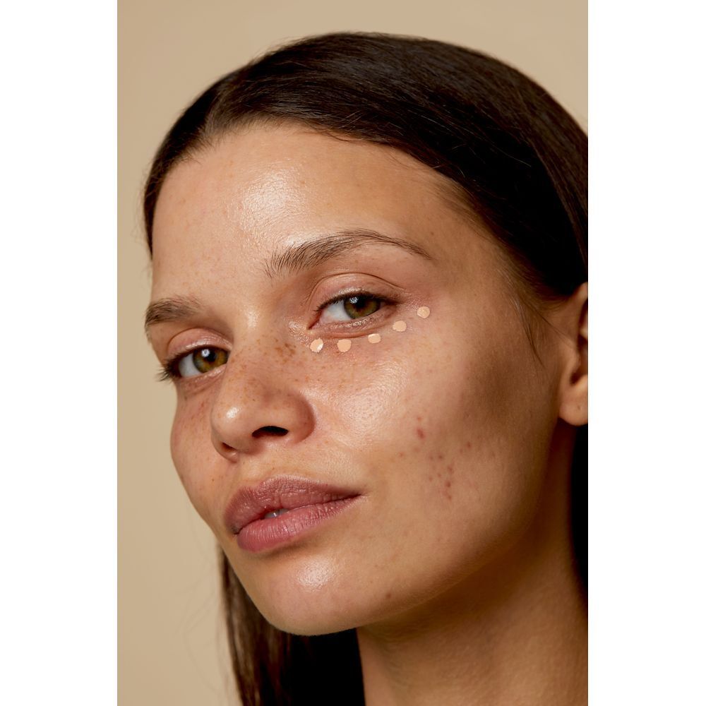 Femme avec points de correcteur sous les yeux. Teint clair, taches de rousseur. Fond beige.