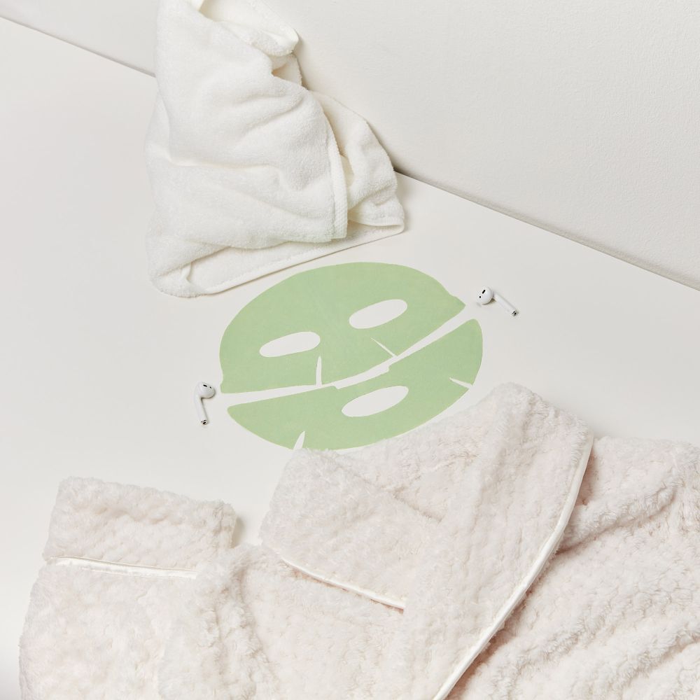 Gesichtsmaske, Handtuch und Bademantel auf weißer Oberfläche. Maske ist hellgrün.