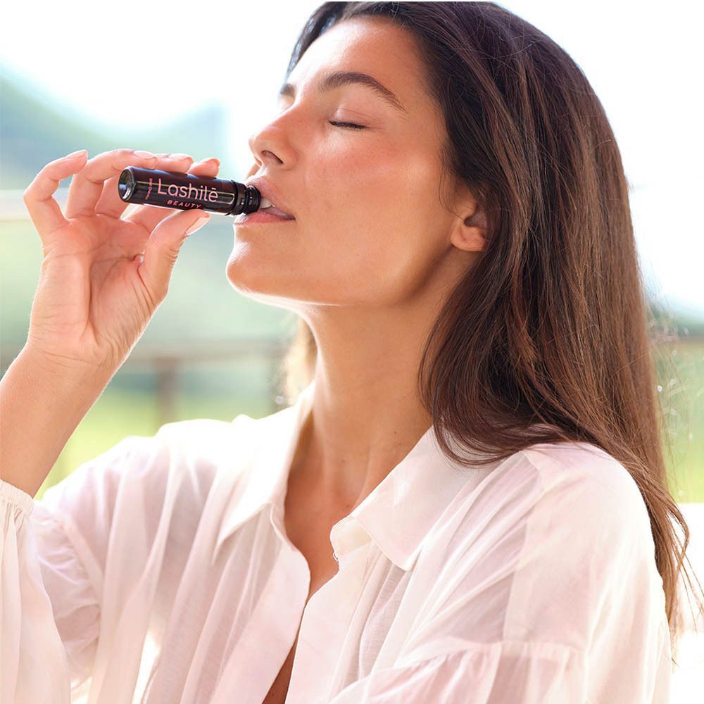 Femme buvant un shot noir Lashilé Beauty. Texte : Good Hair Shots. Fond : fruits.