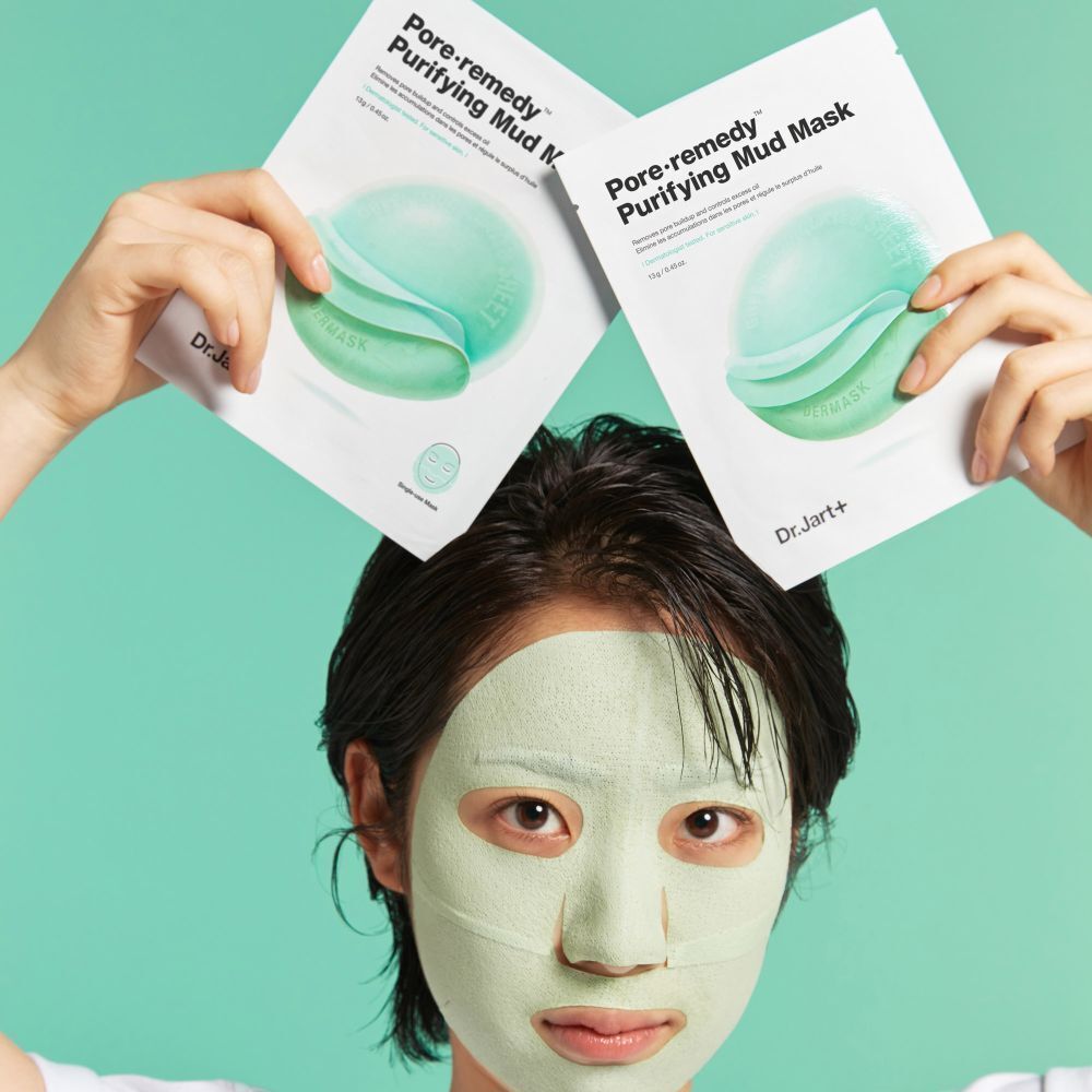 Eine Person mit einer grünen Gesichtsmaske, die zwei Dr.Jart+ Pore·remedy Purifying Mud Mask Päckchen hochhält.