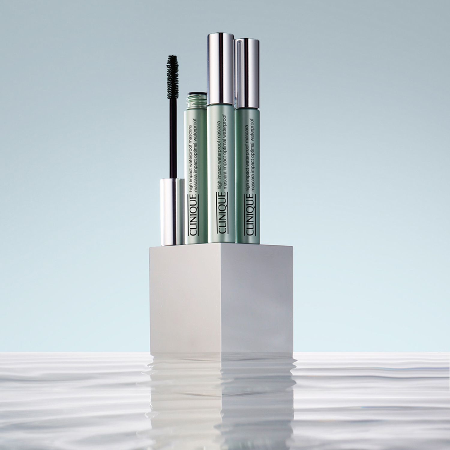 Drei CLINIQUE High Impact Waterproof Mascara-Tuben auf einem weißen Podest. Hintergrund: Wasser mit Reflexionen.