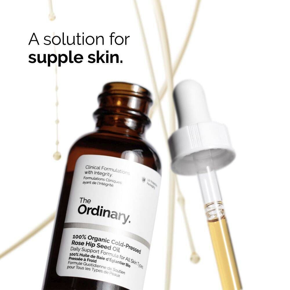 Eine Flasche The Ordinary 100% Bio-Hagebuttenkernöl mit Pipette, umgeben von goldenen Tropfen.