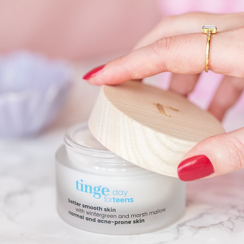 Ein Tiegel mit hellem Holzdeckel, der von einer Hand geöffnet wird. Aufschrift: Tinge day for teens, better smooth skin, mit Wintergrün und Eibisch, für normale und zu Akne neigende Haut.
