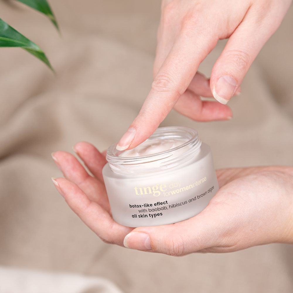 Hände halten Tiegel Crème de Jour Intense. Weißes Glas, Holzdeckel. Aufschrift: Tinge, botox-like effect, für alle Hauttypen.