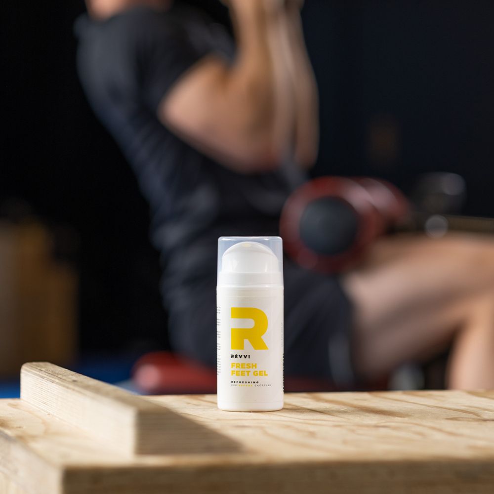 Produit sur bois. Personne s'entraînant en arrière-plan. Flacon blanc avec "R" jaune. Texte : RÉVVI FRESH FEET GEL.
