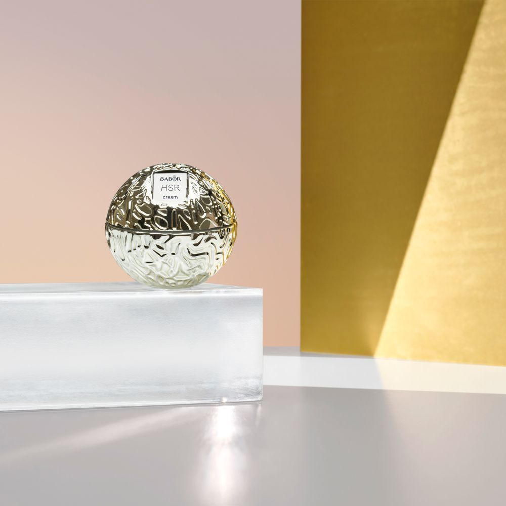 Goldfarbener Cremetiegel mit Babor HSR cream auf einer Glasplatte. Hintergrund: gold und rosa.