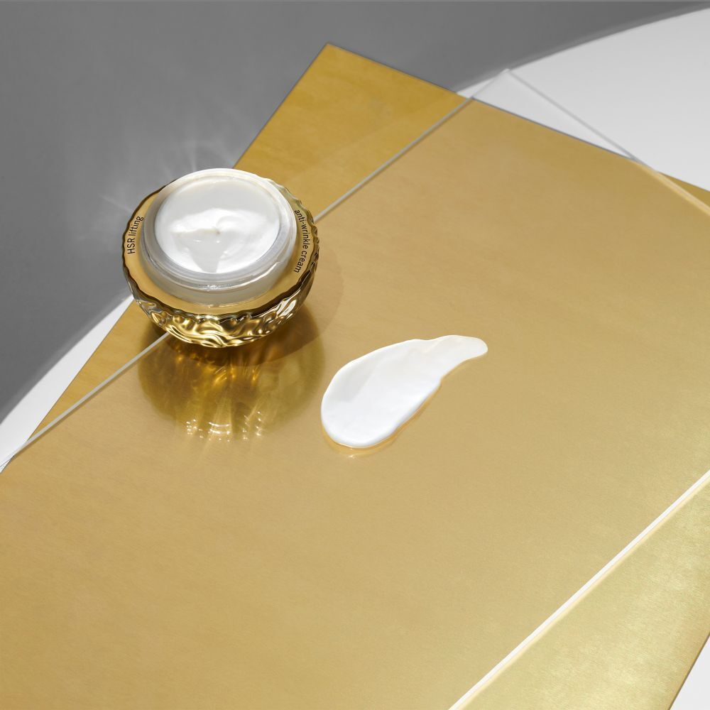 Geöffneter goldfarbener Cremetiegel mit Creme daneben. Hintergrund: goldfarben.